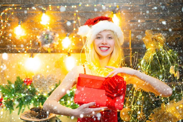 Mooie jonge vrouw in Santa Claus kleding over kerst versierd interieur achtergrond met een geschenk of cadeau. Wintershoppingconcept. — Stockfoto
