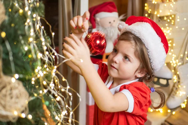 Kleines Mädchen im Weihnachtskleid, das den Weihnachtsbaum mit Kugeln schmückt. Weihnachtskind schmückt Weihnachtsbaum mit Christbaumkugel. — Stockfoto