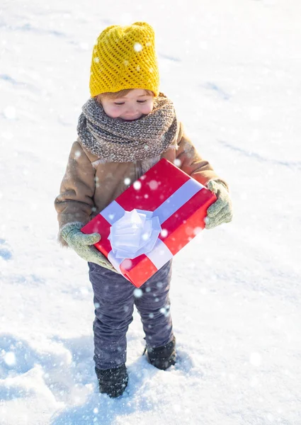 小さな男の子は冬服の帽子とスカーフを閉じます。お正月と陽気なクリスマス。冬休みのコンセプト。素敵な休日をお過ごしください。幸せな冬の子供は贈り物弓雪の背景を保持します。キッド — ストック写真