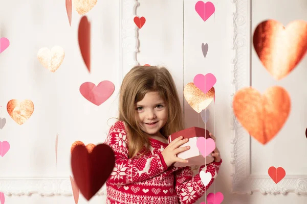 Dia dos Namorados. Pequena menina boneca bonito em camisola de inverno vermelho com flocos de neve e veados segurando um presente caixa dos namorados. A grinalda do coração. Fazer amor e felicidade . — Fotografia de Stock