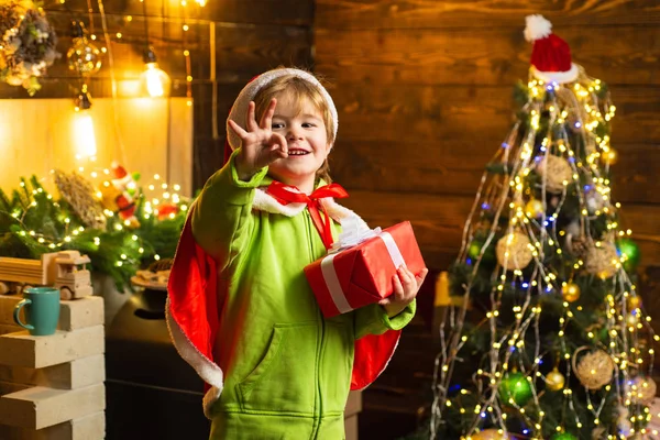 Noel ağacının yanındaki mutlu çocuk elinde Noel hediyesiyle kameraya bakıyor. Küçük çocuk Noel Baba kıyafeti giyiyor.. — Stok fotoğraf