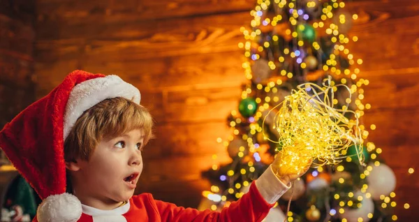 Little kid is playing with Christmas light on Christmas tree background. Happy cute child in Santa hat with present have a Christmas. — Stock Photo, Image