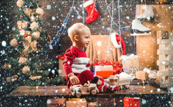 Kind im Schnee. Ein fröhliches kleines Kind in Winterkleidung denkt an den Weihnachtsmann in der Nähe des Weihnachtsbaums. Kinderweihnachten. Winterabend zu Hause. Lächelndes Kind, das im Wohnzimmer hinter dem Weihnachtsbaum hervorlugt — Stockfoto