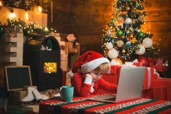 İnternetten çocuklar için Noel alışverişi. Küçük çocuk dizüstü bilgisayarının yanında oturan Noel Baba kıyafetleri giyiyor. Noel konsepti. Şömine arkaplanı. — Stok fotoğraf
