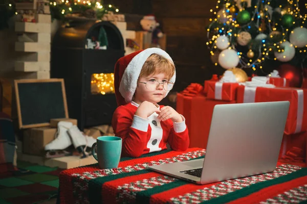 Little kid is wearing Santa clothes sitting by his laptop. Santa helper using notebook. Christmas little boy typing letter to Santa Claus on a computer at home on Christmas background.