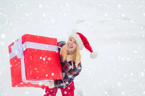 Fille dans la neige. Service de livraison d'hiver. Femme portant un grand cadeau isolé sur fond d'hiver de neige. Portrait de Santa femme avec un énorme cadeau rouge regardant la caméra . — Photo