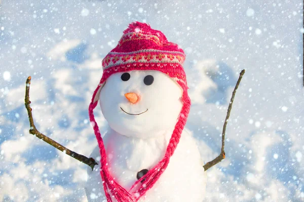 Muñeco de nieve divertido con una zanahoria en lugar de una nariz y en un sombrero de punto caliente en un prado cubierto de nieve sobre un fondo de nieve borrosa. La mañana antes de Navidad. Tarjeta de felicitación de Año Nuevo. Muñeco de nieve en nieve . — Foto de Stock