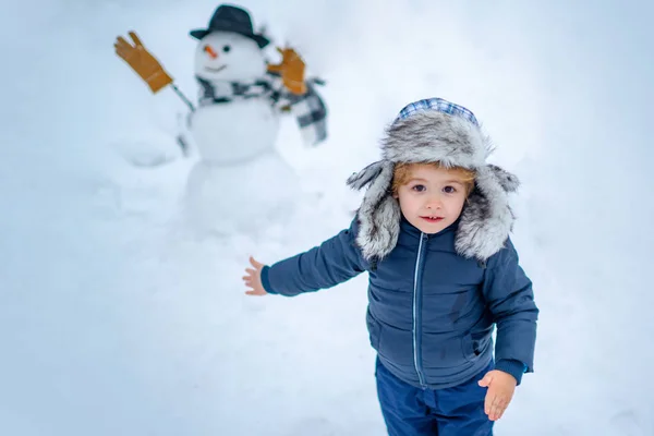 Kleiner Junge spielt mit Schneemann im Winterpark. Winterporträt des niedlichen Kindes im Schneegarten. Menschen im Schnee. glückliches Kind spielt mit einem Schneemann auf einem verschneiten Winterspaziergang. — Stockfoto