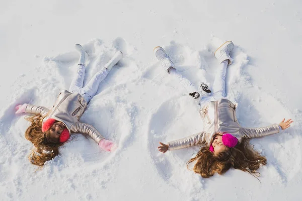 Two Girls on a snow angel shows. Smiling children lying on snow with copy space. Children playing and making a snow angel in the snow. Top view. — Stock Photo, Image