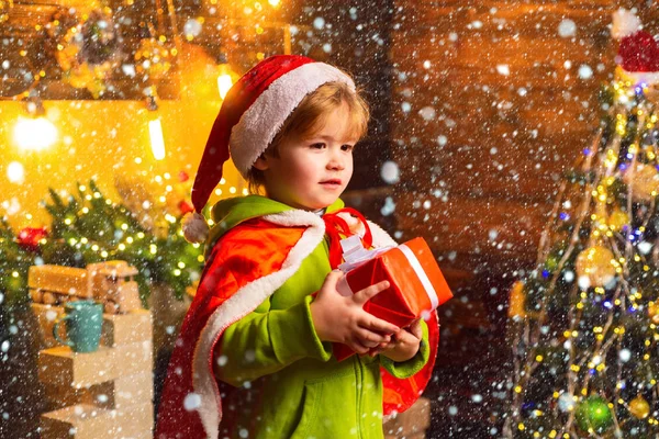 Um miúdo na neve. Um rapazinho feliz junto à árvore de Natal com o seu presente de Natal. O miúdo está a usar roupa de Pai Natal. Conceito de Natal . — Fotografia de Stock