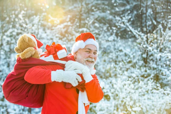 サンタの雪のシーンに対する贈り物の袋を運ぶ。冬のクリスマスグリーティングカード。サンタクロースは白い自然の背景に贈り物の巨大な袋を引く. — ストック写真