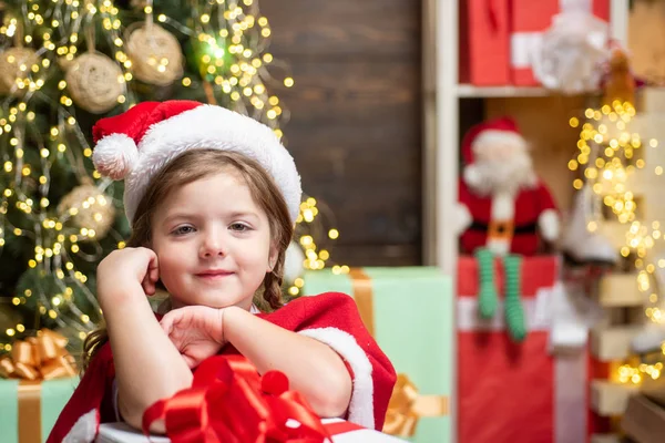 Kind met een kerstcadeau op houten huisachtergrond. Gelukkig kind dat kerstboom versiert. Portret van Santa kind met cadeau op zoek naar camera. — Stockfoto