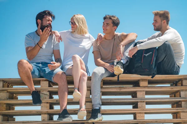 Company of friends students meeting after classes. Charming young bearded man expressively tells his friends an interesting history. Best friends sitting on bench at sky background.