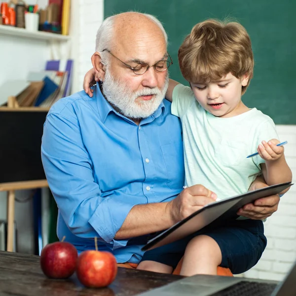 Leraar en leerling leren samen op school. Grootvader praat met kleinzoon. Wereldlerarendag. Kind met oude leraar leren in de klas op de achtergrond van schoolbord. — Stockfoto