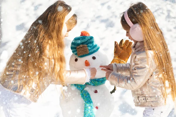 Criança feliz entranhando com um boneco de neve em um passeio de inverno nevado. Tornando boneco de neve e inverno divertido para as crianças. Duas garotinhas brincando na neve no inverno . — Fotografia de Stock