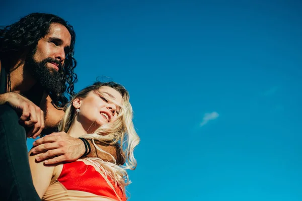 Couple enjoying their strong relations and love. Strong handsome bearded man embracing his young sexy woman in elegant red dress at clear summer blue sky background.