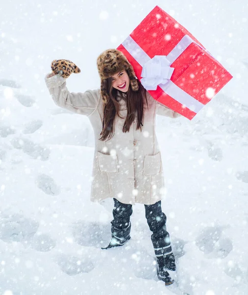 Femme de Noël drôle et une grande boîte cadeau. Femmes de service avec grand coffret cadeau. Femme heureuse debout sur fond de neige blanche et tenant une boîte cadeau rouge géant avec les deux mains . — Photo
