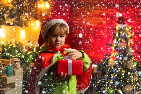 Niedliches Kind öffnet ein Weihnachtsgeschenk. Fröhlicher kleiner Junge als Weihnachtsmann verkleidet. Junge mit Weihnachtsmütze hilft bei Weihnachtsgeschenk in roter Schachtel. — Stockfoto