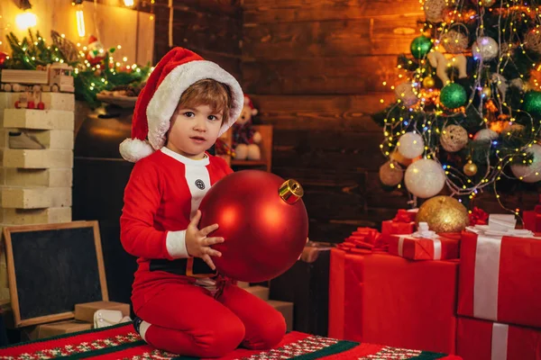 El chico lleva ropa de Santa Claus, decorando el árbol de Navidad. Concepto de Navidad. Niño en Santa hat hold bauble . — Foto de Stock