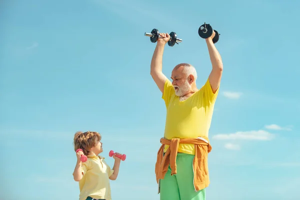 A boldog kisfiú nagyapával súlyzókkal gyakorol. Sportoló nagyapa és egészséges gyerek súlyzós testmozgással. Aktív család élvezze a sport és a fitness. — Stock Fotó