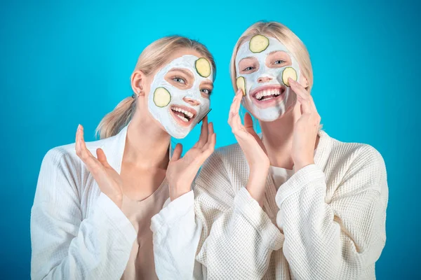 Jonge gelukkige zussen doen gezichtsmasker met plakjes komkommer op blauwe achtergrond. Huidverzorging en -behandeling, spa, natuurschoon en kosmetologie. — Stockfoto