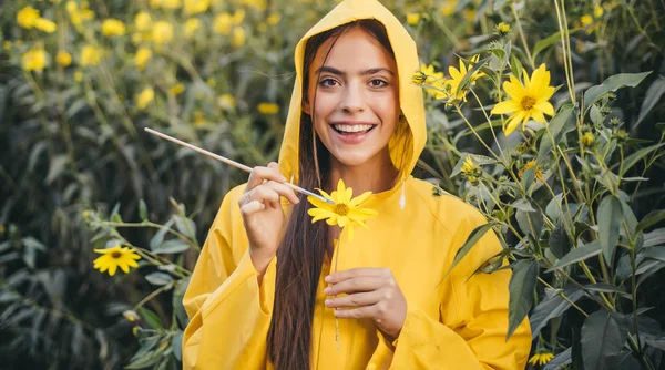 美丽的微笑的年轻女子画黄色的花。 一个笑容可亲的姑娘,戴着黄色的帽衫,用刷子刷着黄色的花. 花儿绽放与自然爱的概念. — 图库照片