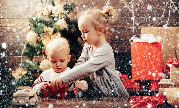 Eröffnungsgeschenke zu Weihnachten und Neujahr. Weihnachtskarte. Porträt Kind mit Geschenk auf Holzgrund. Lächelndes Kind lugt hinter Weihnachtsbaum im Wohnzimmer hervor. — Stockfoto
