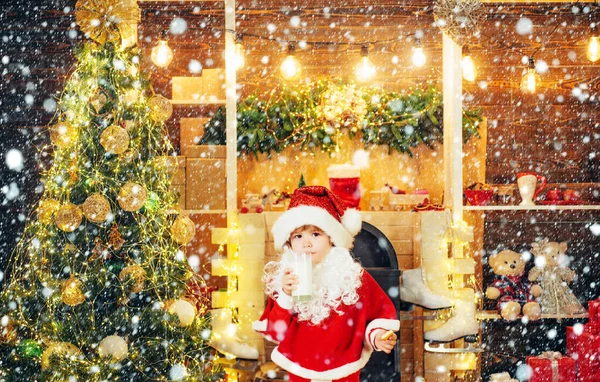 Portret van een kind met een baard in kerstmankostuum. Vrolijk kerstfeest en gelukkig nieuwjaar. Kerstman koekje en glas melk op kerstboom achtergrond. Stockafbeelding