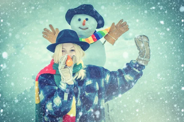 Chica divertida haciendo muñeco de nieve con nariz de zanahoria divertida. Muñeco de nieve divertido con una zanahoria en lugar de una nariz y en un sombrero de punto caliente en un prado cubierto de nieve sobre un fondo de nieve borrosa . — Foto de Stock