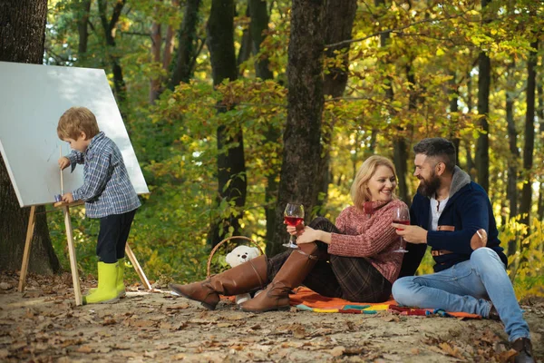 Piccolo figlio intelligente che disegna la sua famiglia dando momenti gioiosi a mamma e papà. Genitori che scelgono l'anniversario di matrimonio. Un picnic in famiglia. Madre, padre, figlio che disegnano insieme a Green Park. Famiglia — Foto Stock