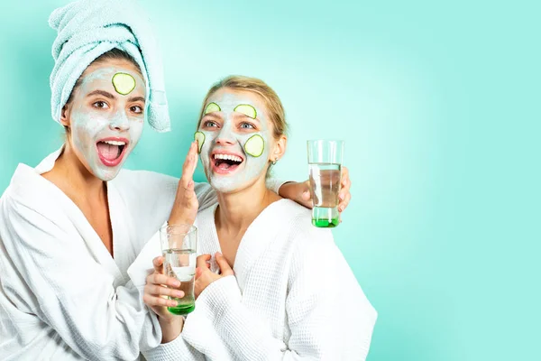 Happy healthy beautiful girls with face mask and slices of cucumber smile and make a skincare. Drink glass of water and laugh. Healthy and wellness concept. — Stock Photo, Image