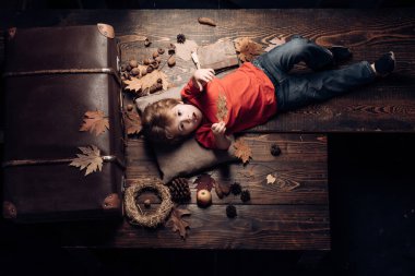 Çocuklar için tüm sonbahar kıyafetlerinde en büyük indirim. Sarışın, küçük bir çocuk, sonbahar yapraklarında, midesinde yaprak, ahşap zeminde yatıyor. Sonbaharda oynayan çocuk.