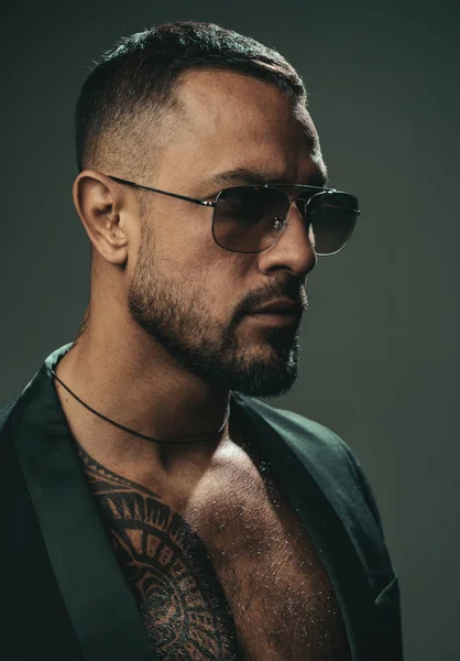 Sexy barbudo tatuado homem vestindo óculos de sol e foto de estúdio rosto dramático. Homem musculoso forte representante do estilo masculino e da moda. Conceito de estilo de vida de luxo . — Fotografia de Stock
