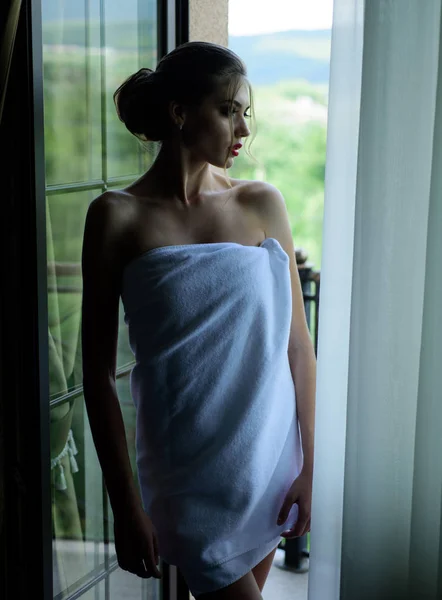 Sinnliche junge Frau in Handtuch gehüllt, nachdem sie unter der Dusche am Fenster stand und nach draußen schaute. schöne Frau mit Luxus-Make-up und Frisur am Fenster Hintergrund. — Stockfoto