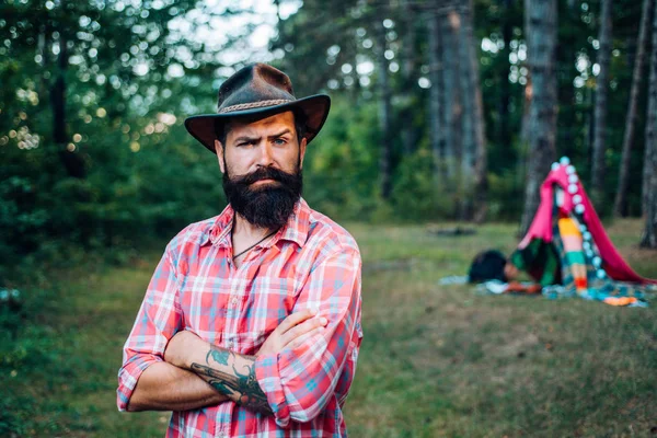 森林里的家伙 夏令营。 长胡子的野蛮牛仔 野蛮的黑发男人戴着帽子,在树后. 戴帽子留着胡子的成熟男人. 希普特风格的远足于深林中. — 图库照片