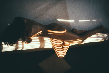 Işık ve gölge oyunu. Kadın vücudunun güzelliği. Güneş panjurlarının gölgesiyle bronzlaşmış ince bir kadın vücudu. Sabahları yatakta yatan baştan çıkarıcı genç bir kadın..