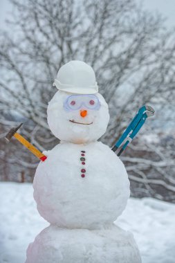 Kar tarlasında iş kasklı komik bir kardan adam. Açık havada el yapımı kardan adam. Kar arkaplanda çekiçli bir kardan adam..