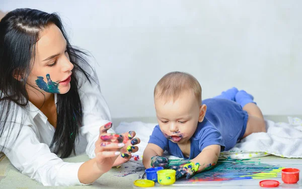 Baby child draws with colored paints hands. Loving mother with her baby. Beautiful baby covered in bright paint. Young mother playing with her newborn son. Family concept.