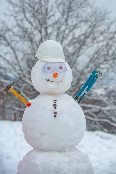 Grappige sneeuwpop in werkhelm op besneeuwd veld. Handgemaakte sneeuwpop in de sneeuw buiten. Sneeuwman werknemer met hamer op sneeuw achtergrond. — Stockfoto