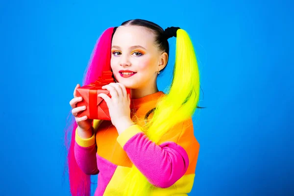 Sevimli doğum günü kızı elinde kırmızı bir kutuyla kameranın mavi arka planına bakıyor. Parti için renkli makyaj ve saç kesimi. — Stok fotoğraf