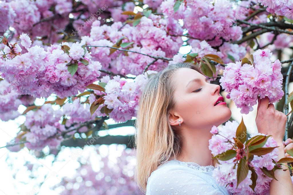 Spring woman fashion concept. Blonde woman with blooming Sakura tree and sunny day. Blooming sakura blossoms flowers close up with blue sky on nature background.