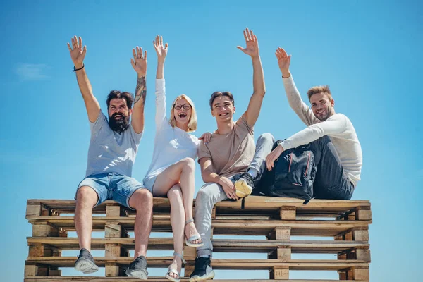 Grupo de amigos alegres estudiantes sentados en el banco y levantando las manos en el fondo azul del verano. Comunicarse con las personas y el concepto de amistad . — Foto de Stock