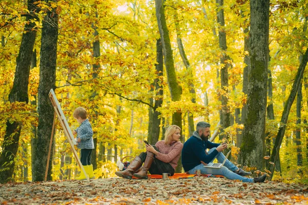 Sunny warm autumnal day at park with family. Wife stalkering a messages of her husband. Little kiddy son paints with brush. Family weekends outside at autumn season concept. — Stock Photo, Image