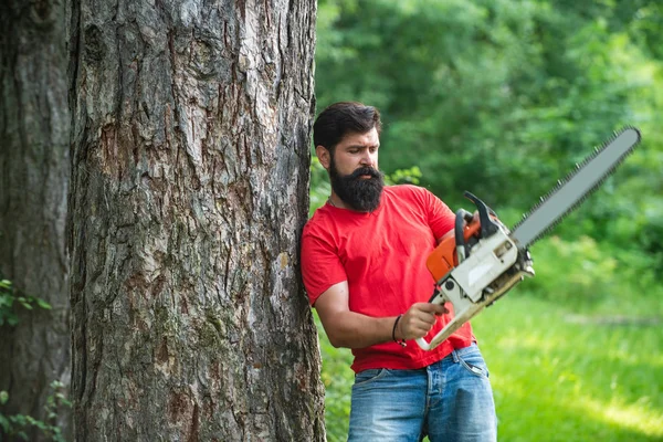 Un bel giovanotto con la barba porta un albero. Operaio boscaiolo che cammina nella foresta con la motosega. Operaio boscaiolo in piedi nella foresta con motosega . — Foto Stock