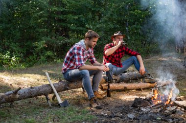 Kamp ateşinin yanında rahatlayan iki erkek sırt çantalı grup. Bir grup genç dışarıda eğleniyor. Piknik arkadaşları. Hafta sonu. Gençler yaz günü parkta piknik yapıp bira içiyorlar..