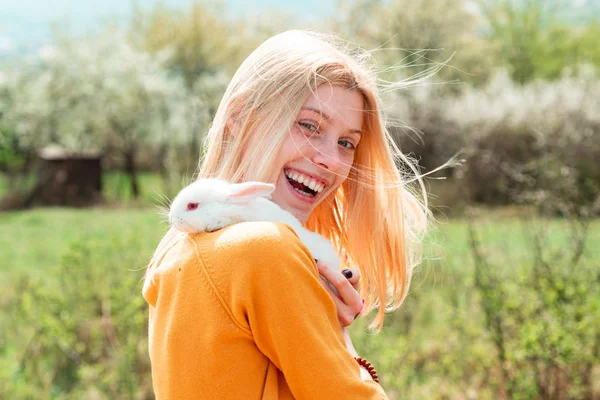Fille drôle tenir lapin sur fond de champ vert. Gros plan de clin d'oeil fille avec lapin lapin. Belle femme tenir lapin blanc . — Photo