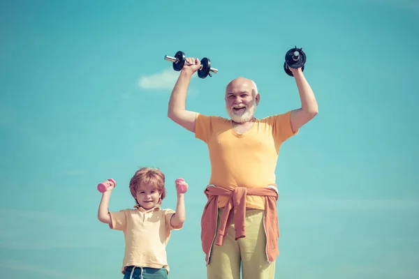 Mosolygó kisfiú és boldog, jóképű öregember, aki súlyzókkal edz. Nagyapa segít a gyereknek súlyzókkal edzeni. Nagyapa és gyermek emelő súlyzó. — Stock Fotó