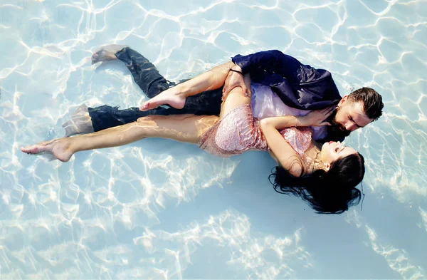 Summer couple cuddles in the pool. Wet evening dresses. Tenderness in clear water. Advertising and commercial design. A bearded man in a jacket and a girl in a dress fell into the water. — Stock Photo, Image