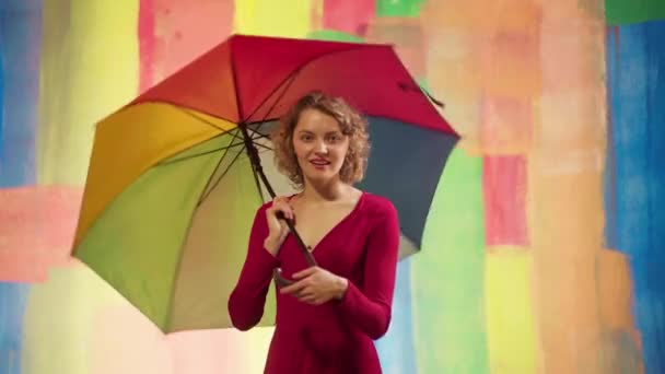Vidám kaukázusi fiatal nő szivárvány esernyővel szórakozás színes stúdió háttér. Vicces érzelmi lány át fényes háttér. — Stock videók