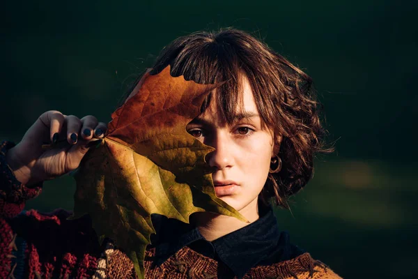 Ambiente de naturaleza otoñal. Primer plano retrato de una guapa morena escondiendo su cara detrás de una hoja amarilla. Hermosa chica sin maquillaje. Muchacha atractiva estudiante con suéter elegante . — Foto de Stock